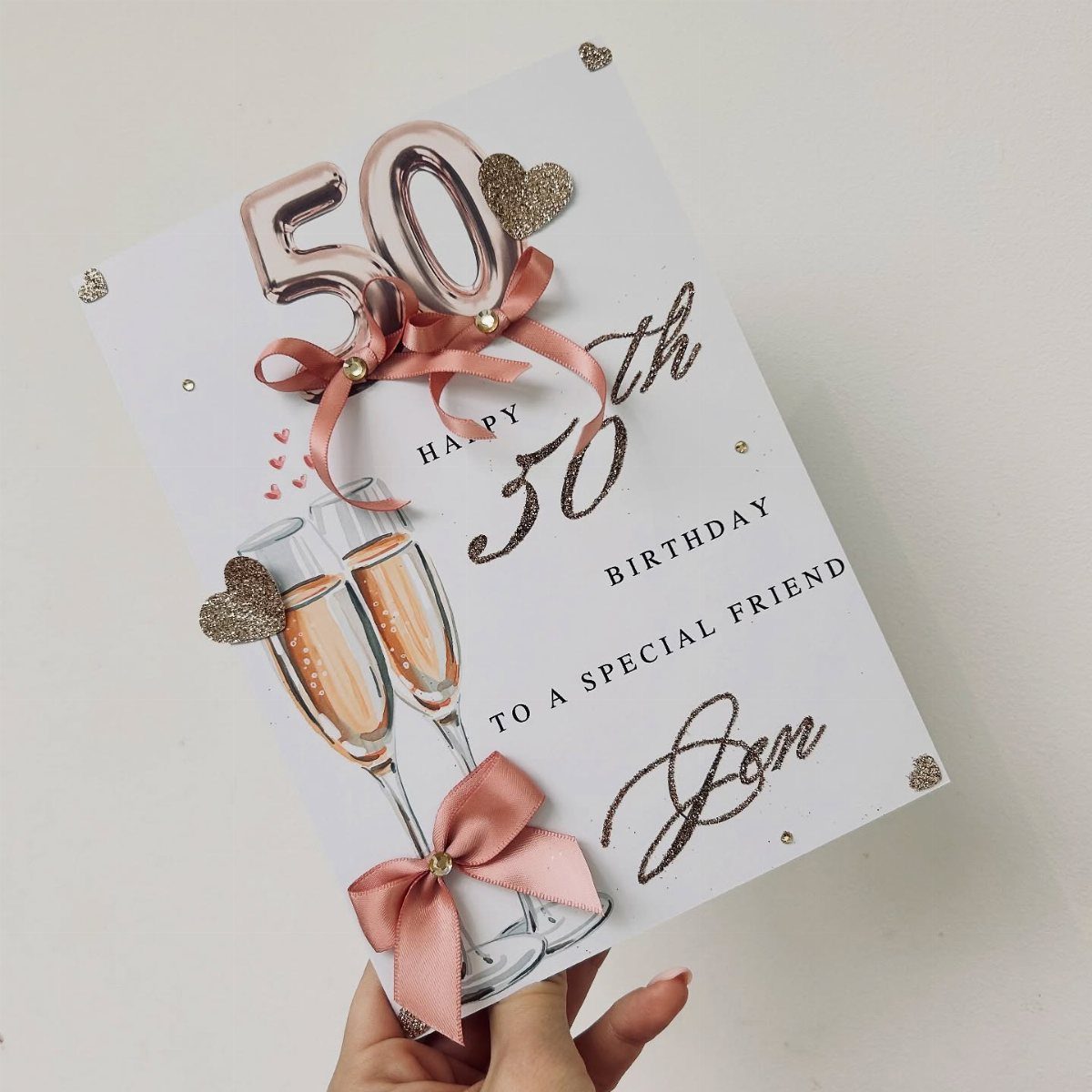 White 50th birthday card with champagne glasses, rose gold balloons, and pink ribbons.