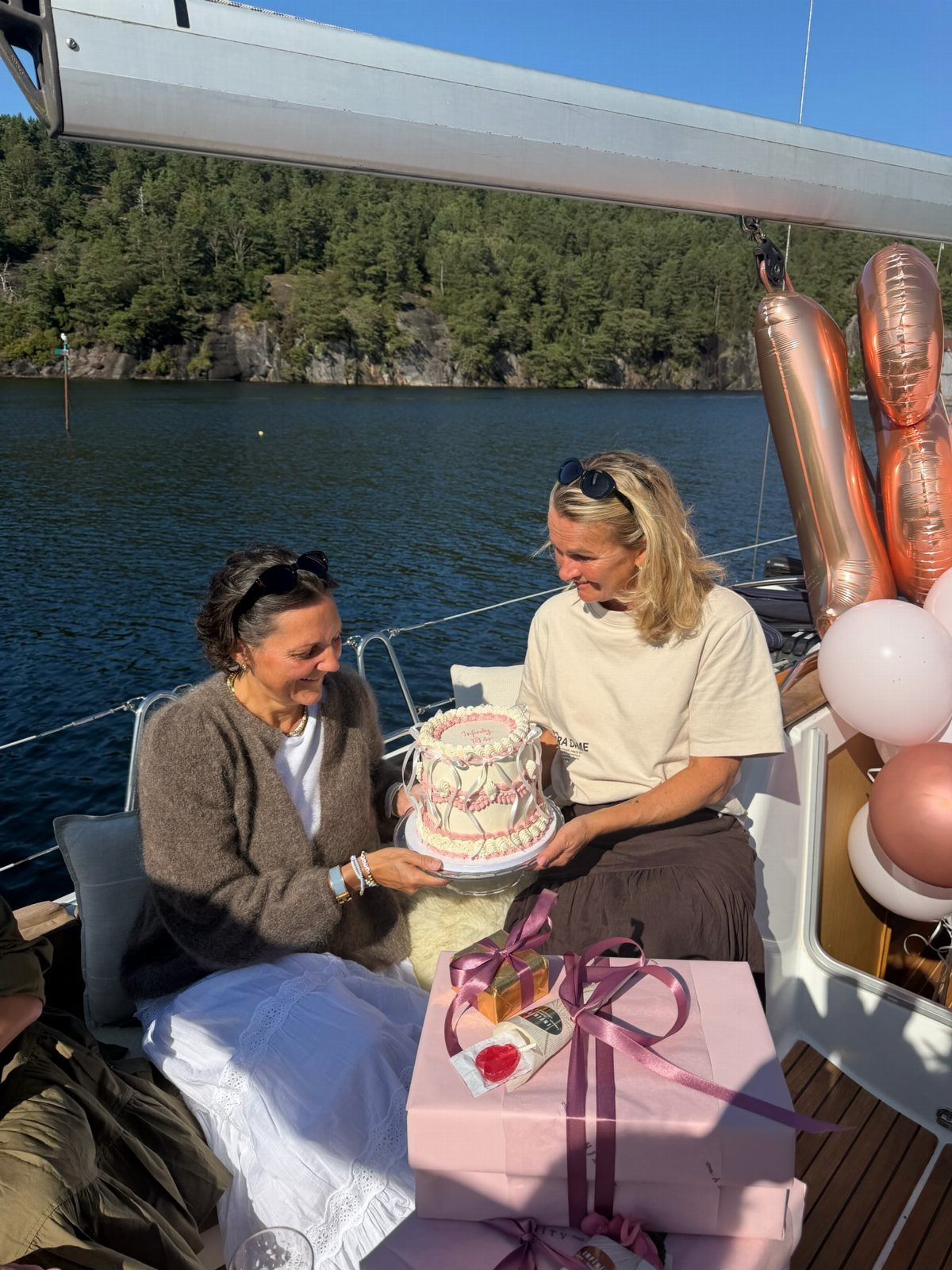 Two women holding a birthday cake on a sailboat deck near a lake.