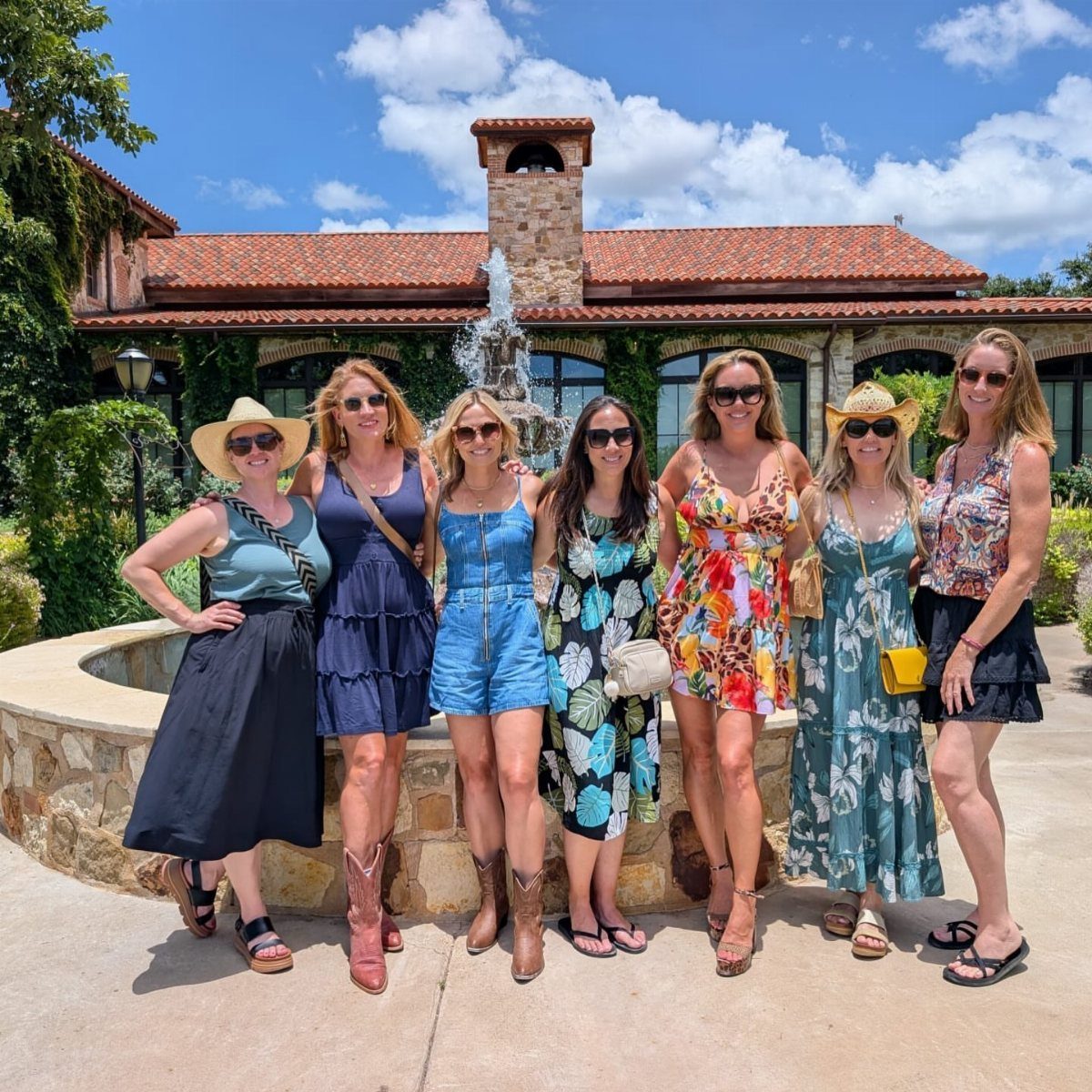 Six women in summer attire posing in front of a fountain and villa.