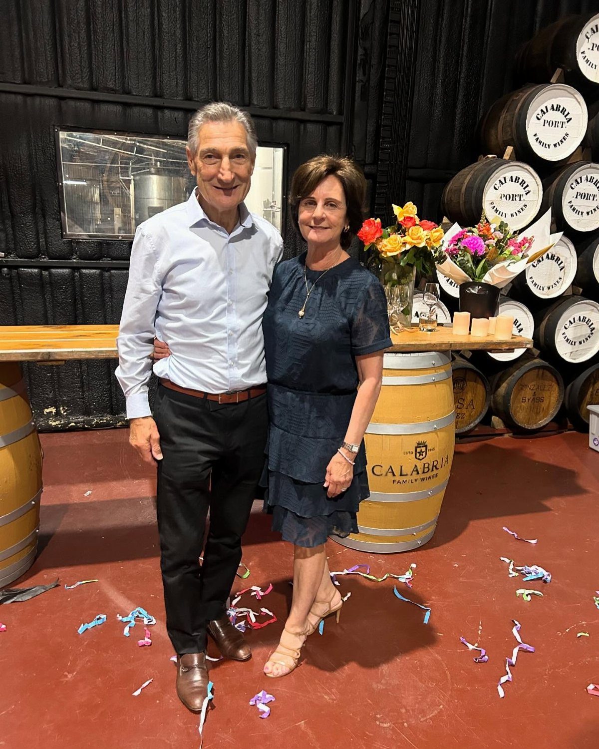 Middle-aged couple posing in a winery surrounded by wooden wine barrels and flowers.