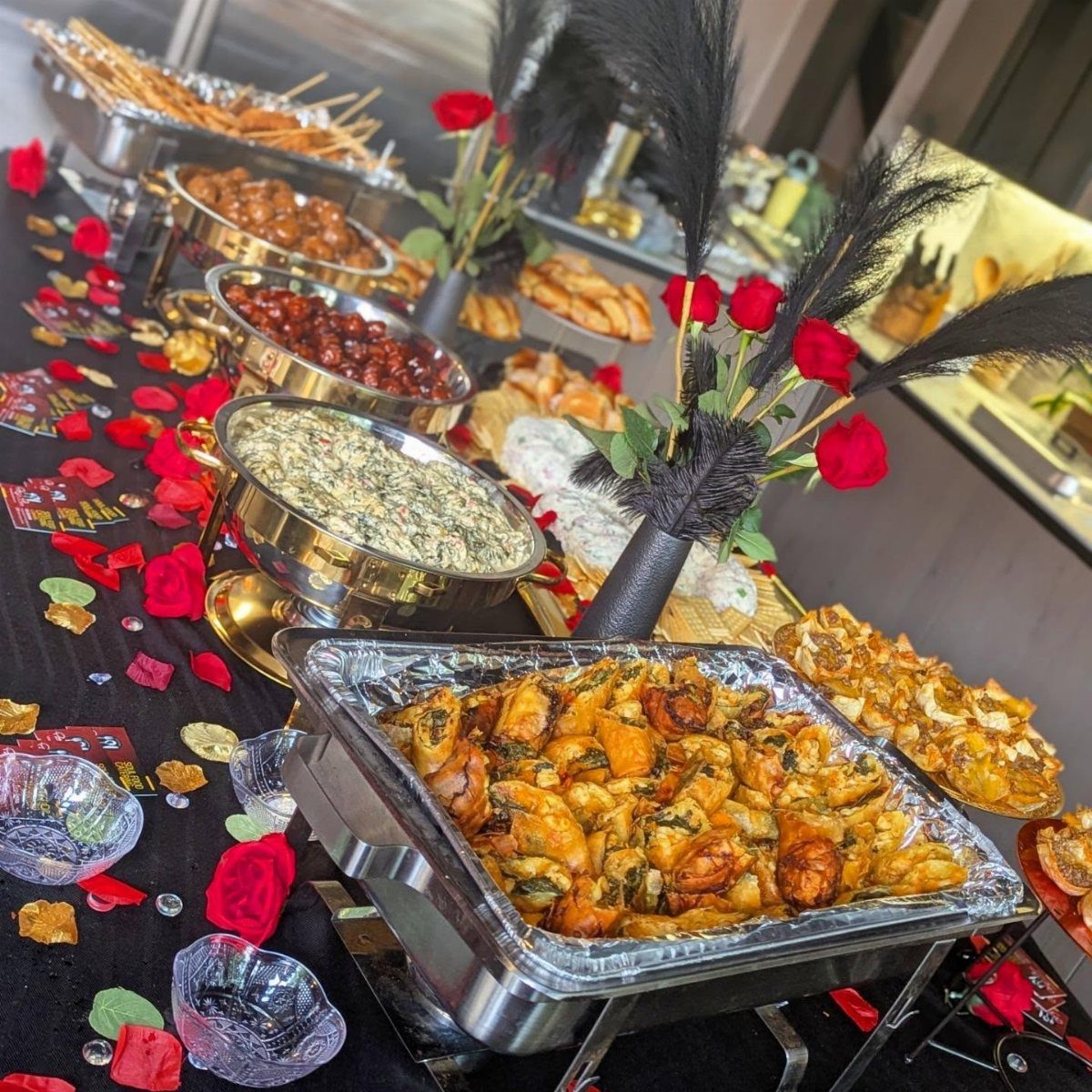 Buffet table with gourmet food, red roses, and black feathers for a celebration.