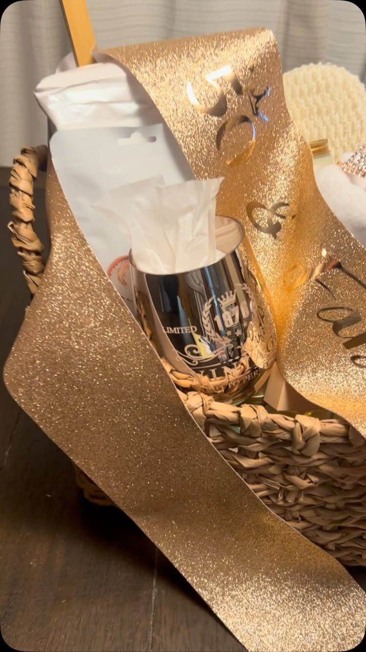 Woven gift basket with white linens, a silver cup, and gold glitter ribbon.