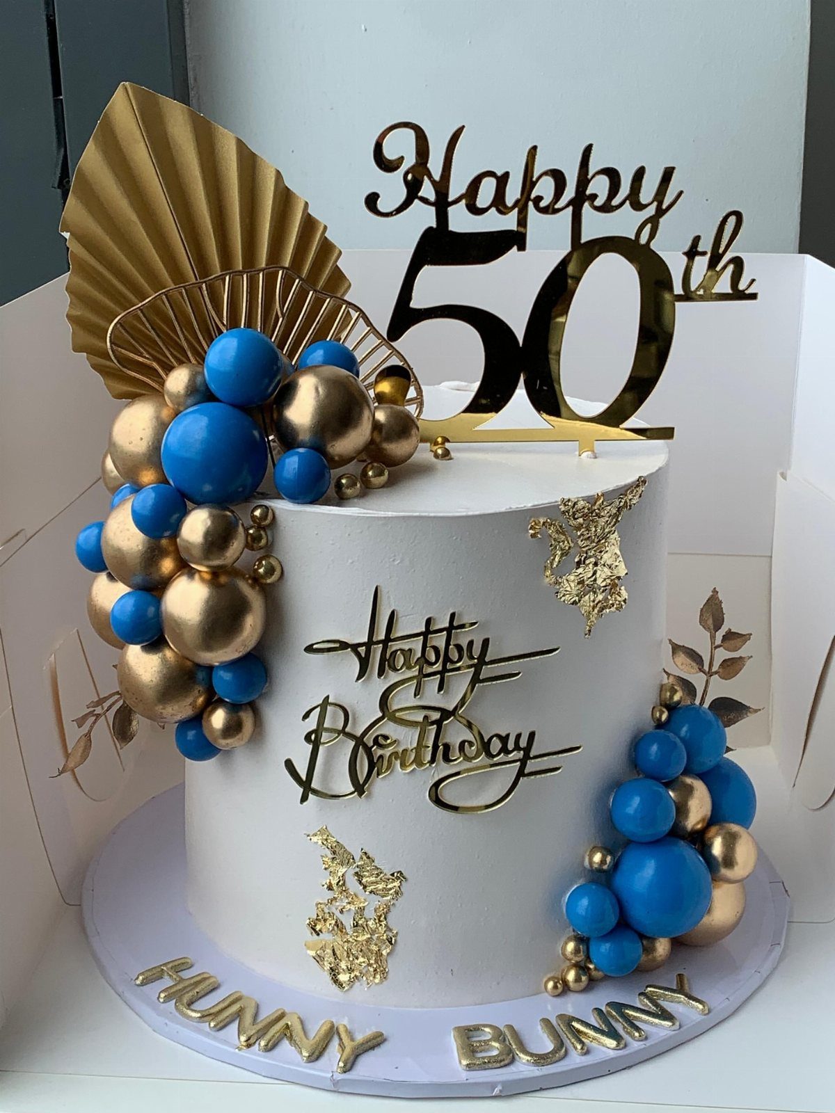 White 50th birthday cake with blue and gold spheres and gold toppers.
