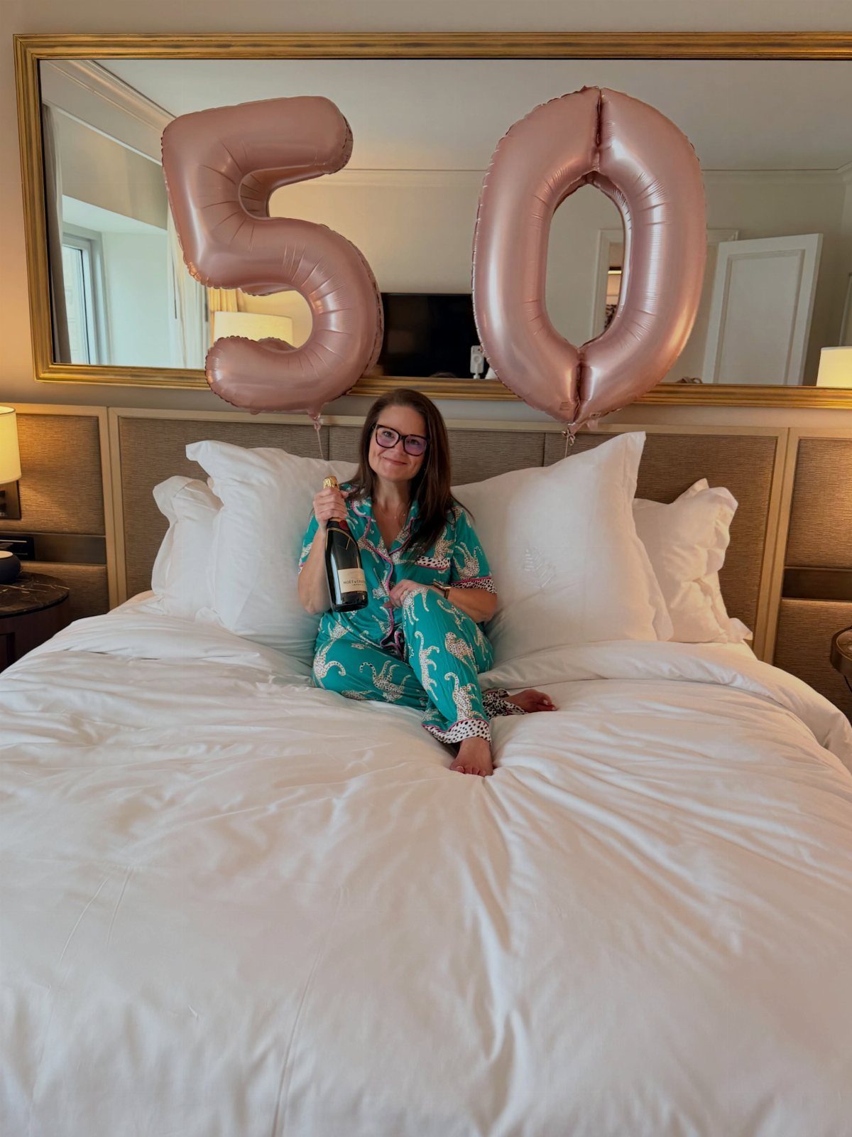 Woman in pajamas sitting on a hotel bed with rose gold 50 balloons.