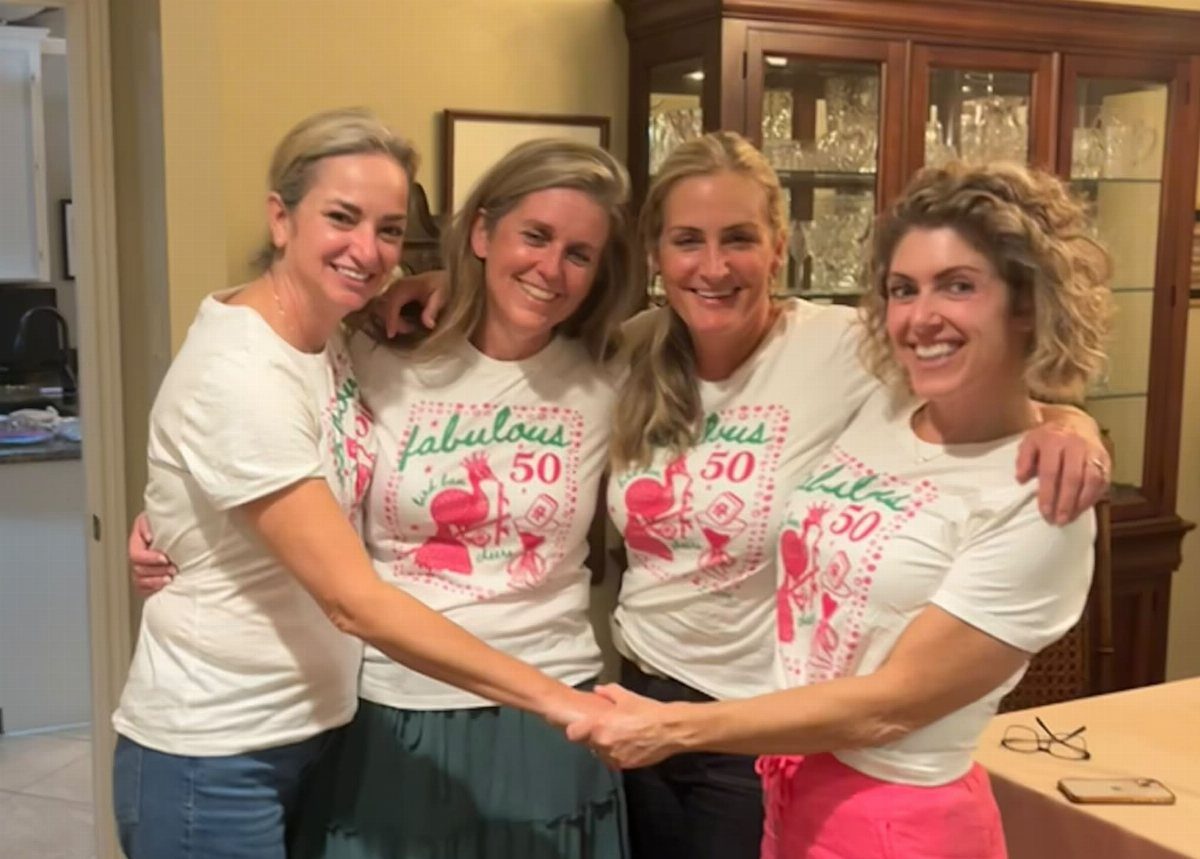 Four women smiling and holding hands, wearing white fabulous 50 birthday t-shirts.