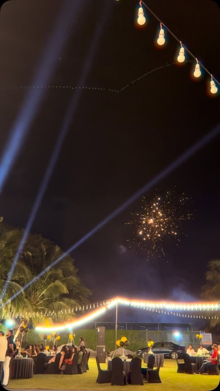 Outdoor nighttime party with guests at tables, string lights, and fireworks in sky