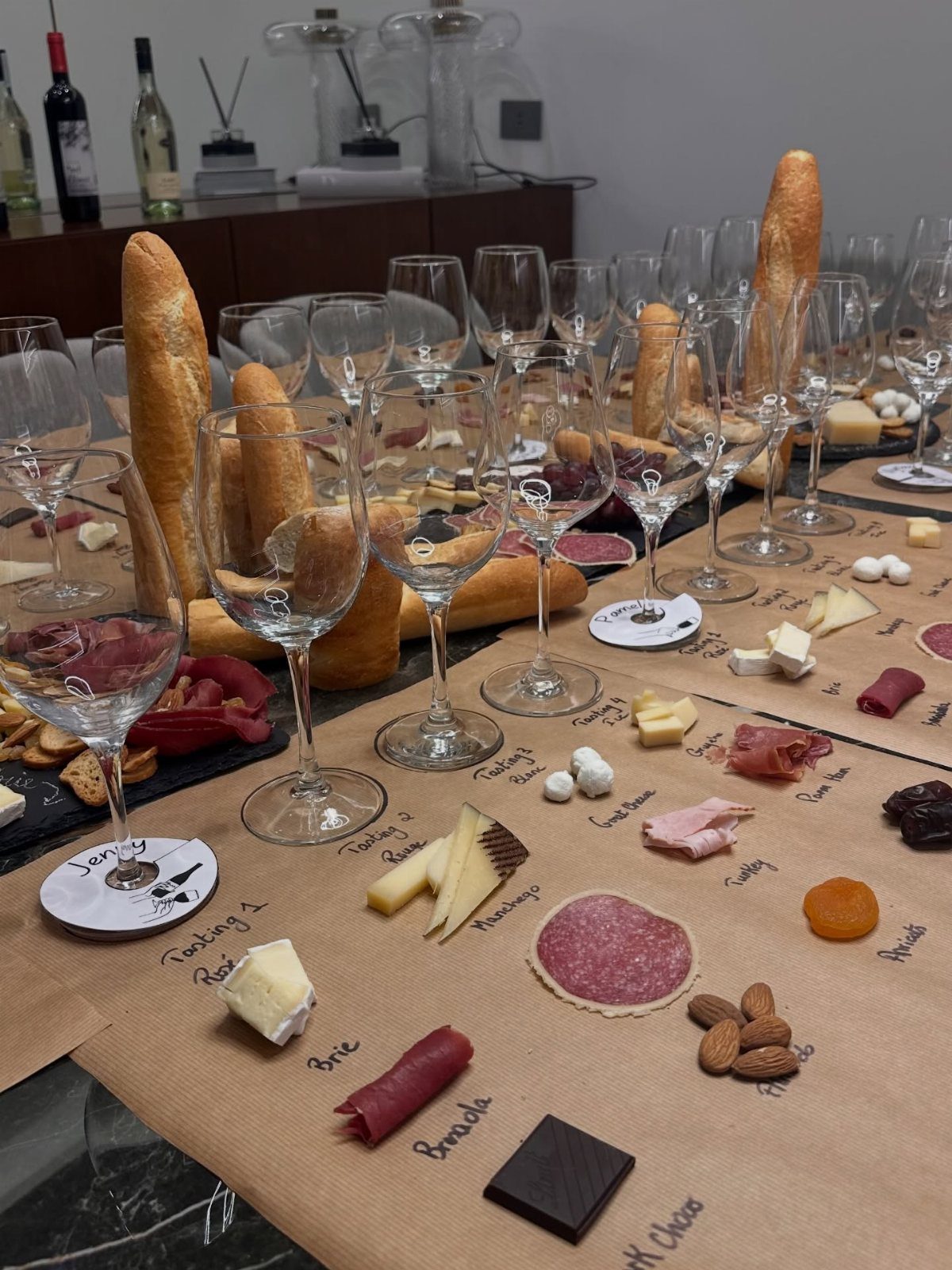 Wine tasting spread with glasses, baguettes, cheeses, and cured meats on brown paper.