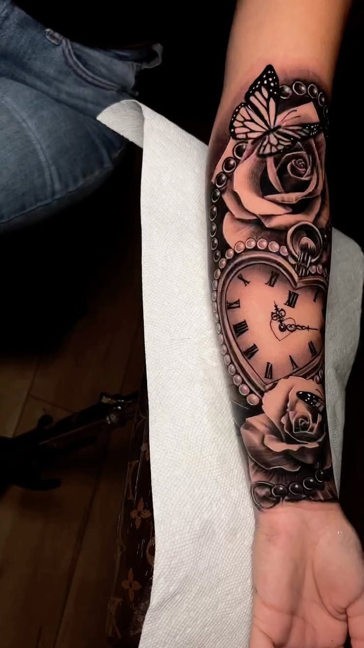 Forearm tattoo featuring a heart-shaped clock, roses, and a butterfly in black ink.