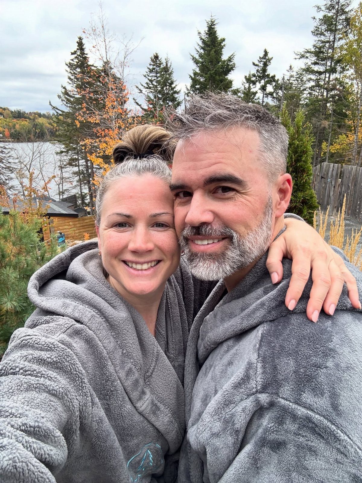 Smiling middle-aged couple in grey bathrobes posing outdoors by a lake and forest.