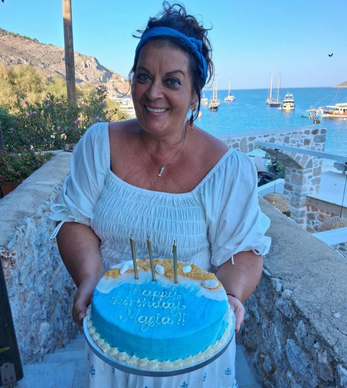 Woman holding a blue birthday cake overlooking a Greek bay with sailboats.