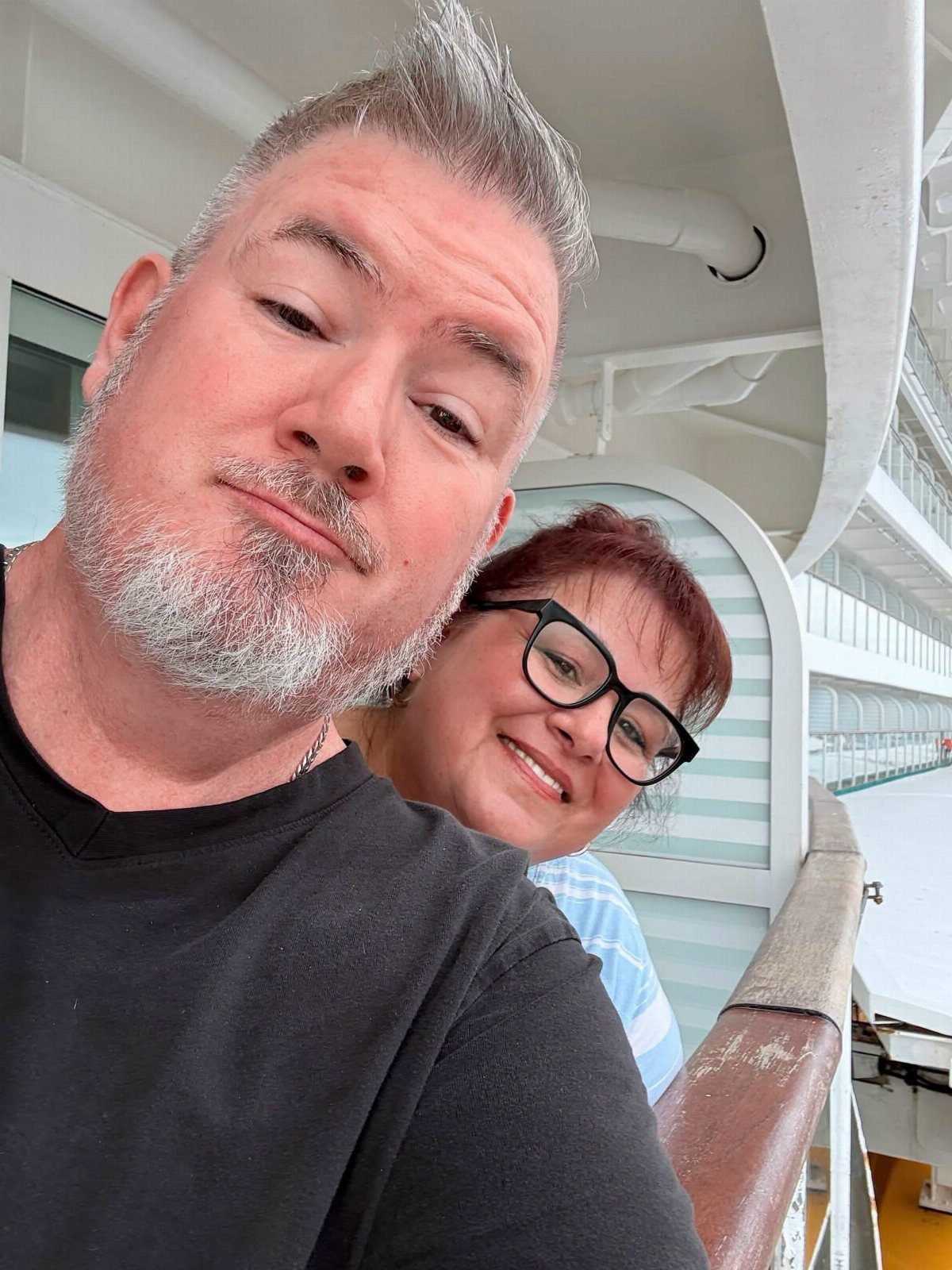Middle-aged couple smiling for a selfie on the white deck of a cruise ship.