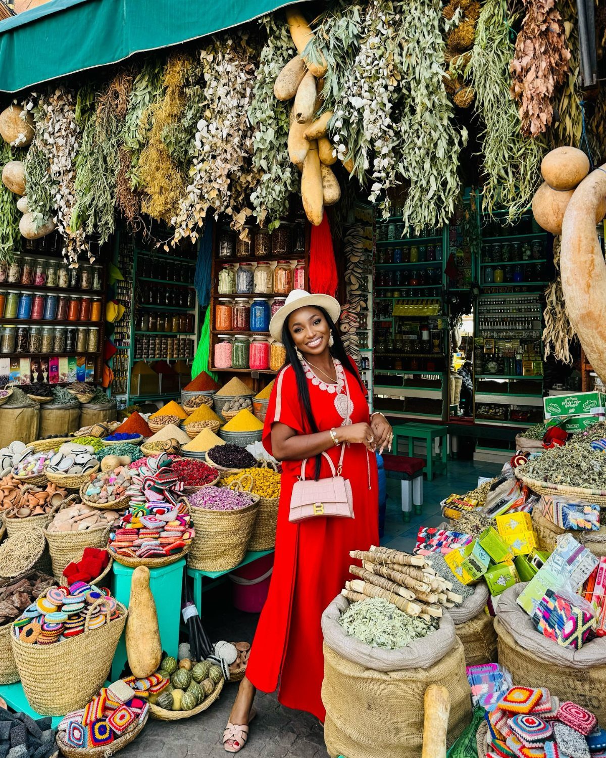 Woman in red dress smiling at a colorful Moroccan spice market with hanging herbs.