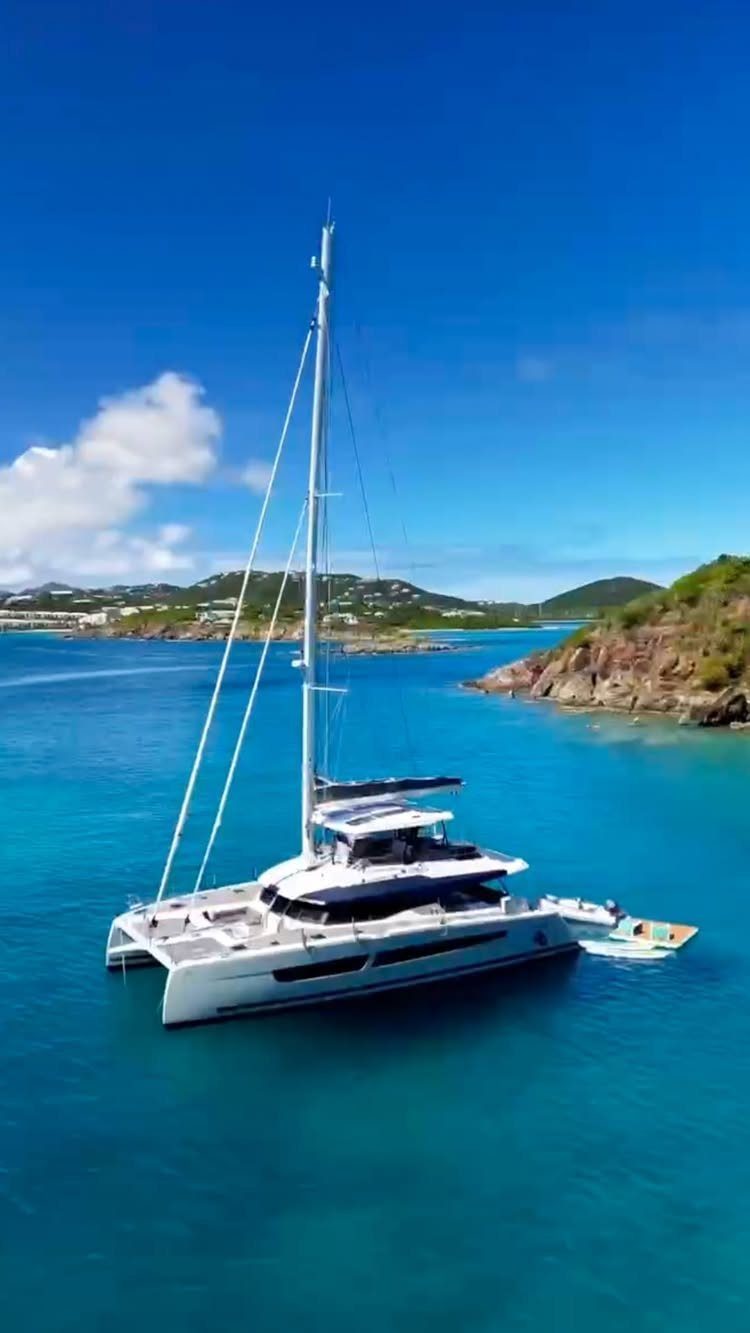 White luxury catamaran sailboat anchored in turquoise tropical waters near green hilly coastlines.