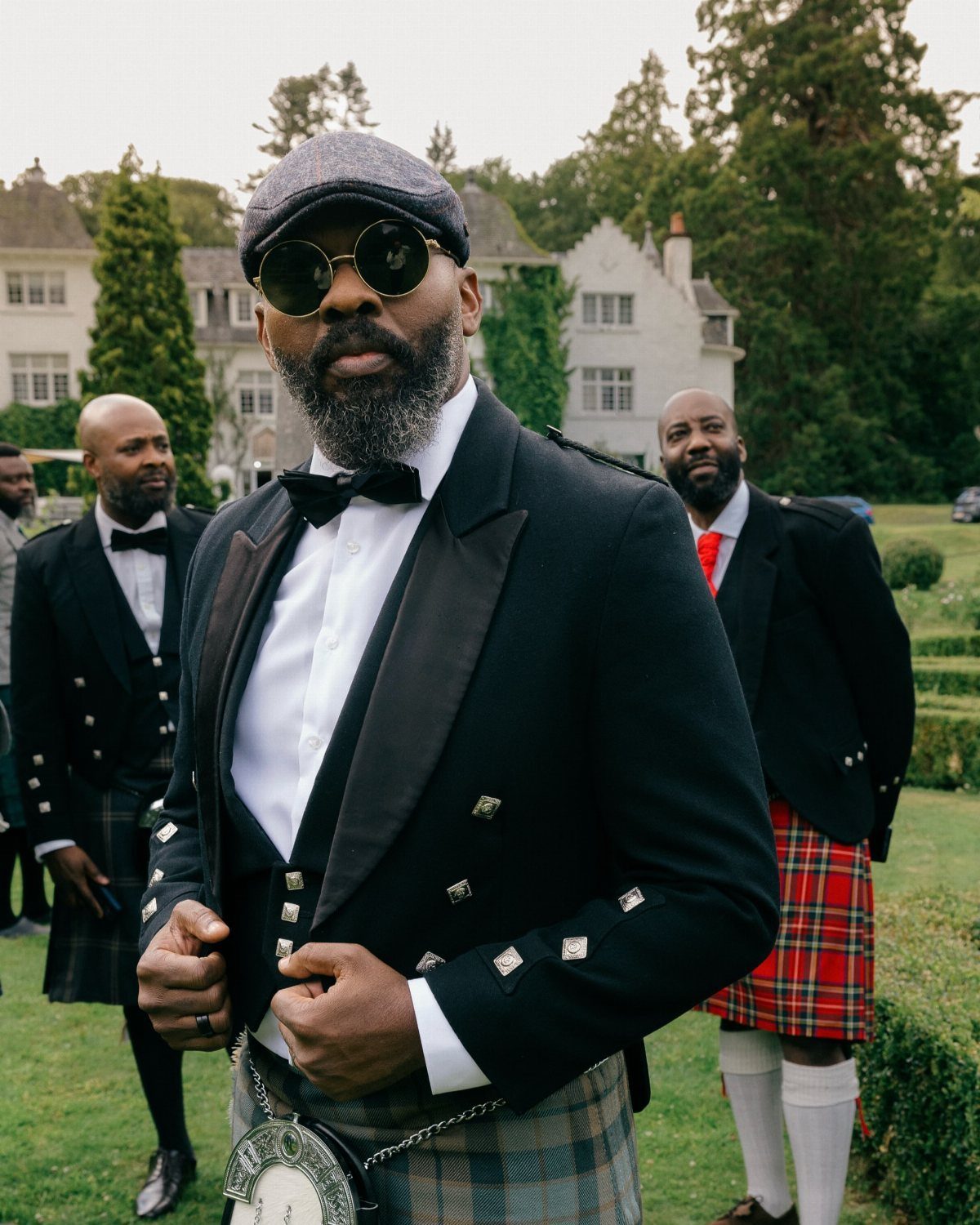 Men in traditional Scottish kilts and formal jackets standing before a white estate.