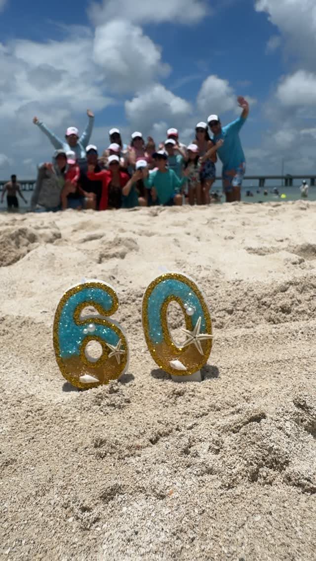 Glittery number 60 with seashells on a beach with people posing behind.