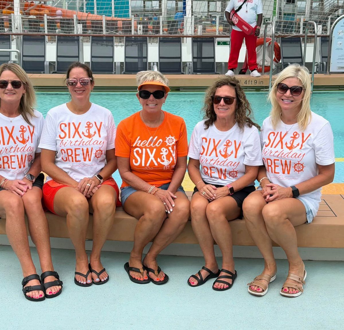 Five women in matching 60th birthday crew t-shirts sitting by a cruise ship pool.
