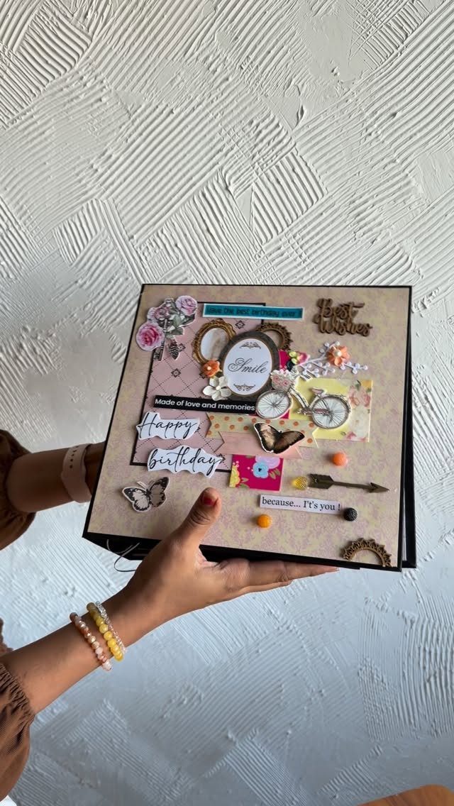 Hands holding a decorated birthday scrapbook page with flowers and a bicycle illustration.