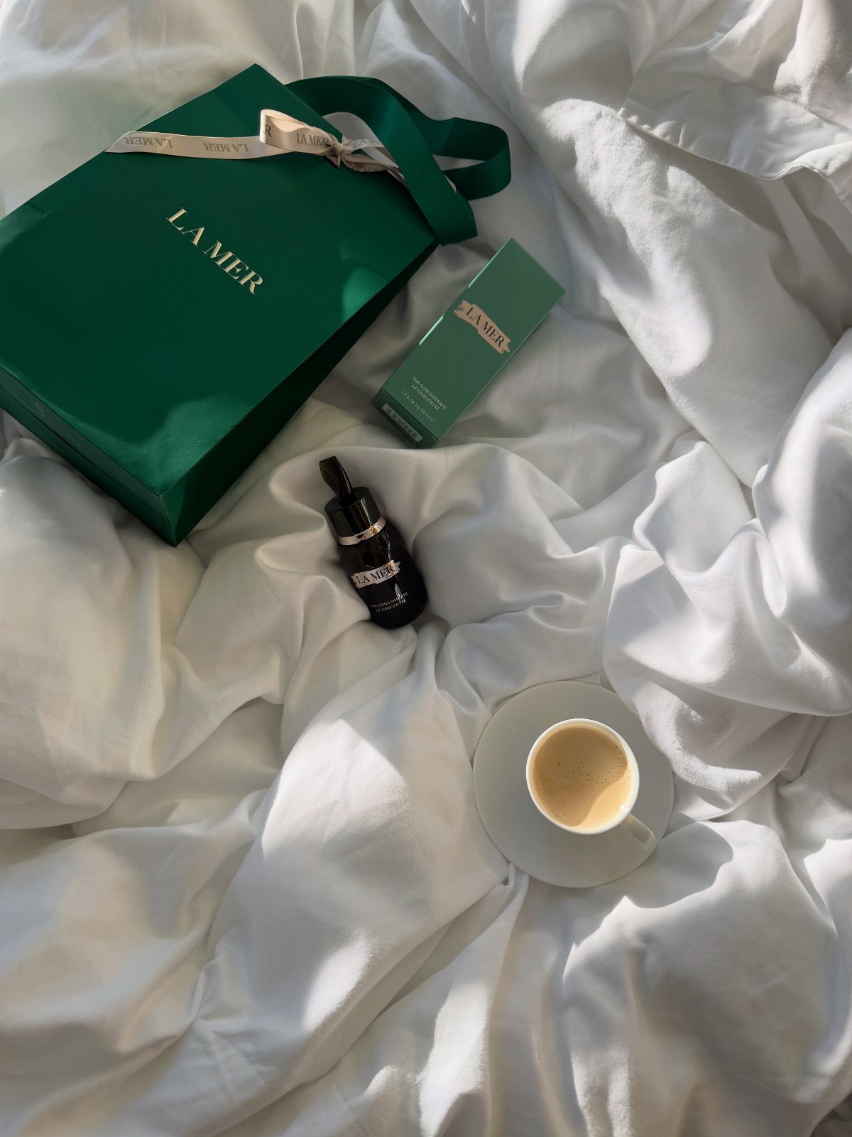 La Mer skincare products and a shopping bag on white sheets with coffee.