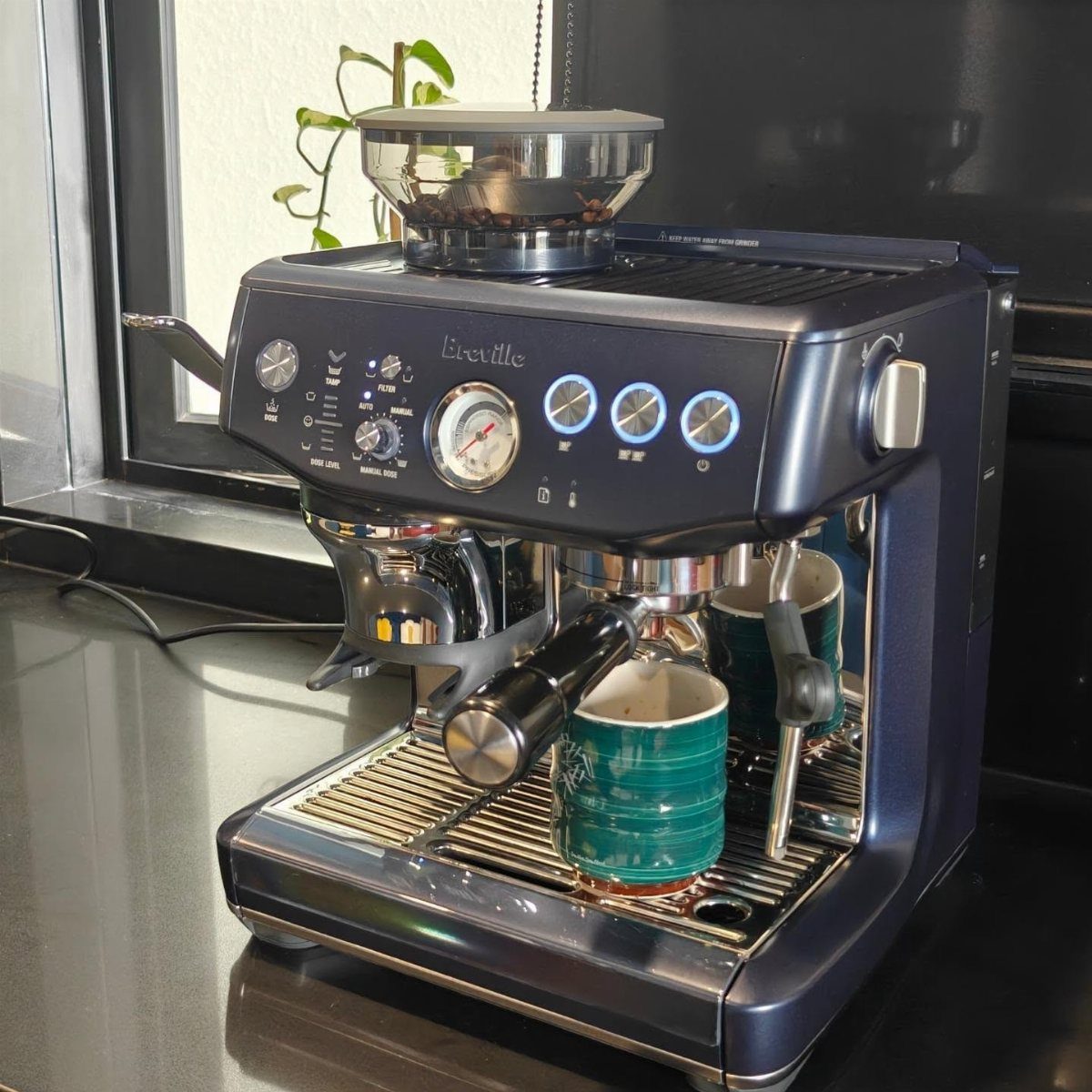 Metallic blue Breville espresso machine with two teal mugs on a dark counter
