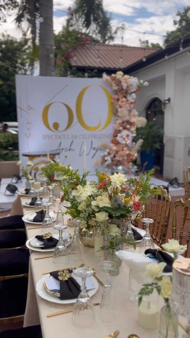 Elegant outdoor 60th birthday party table setting with gold accents and floral backdrop.