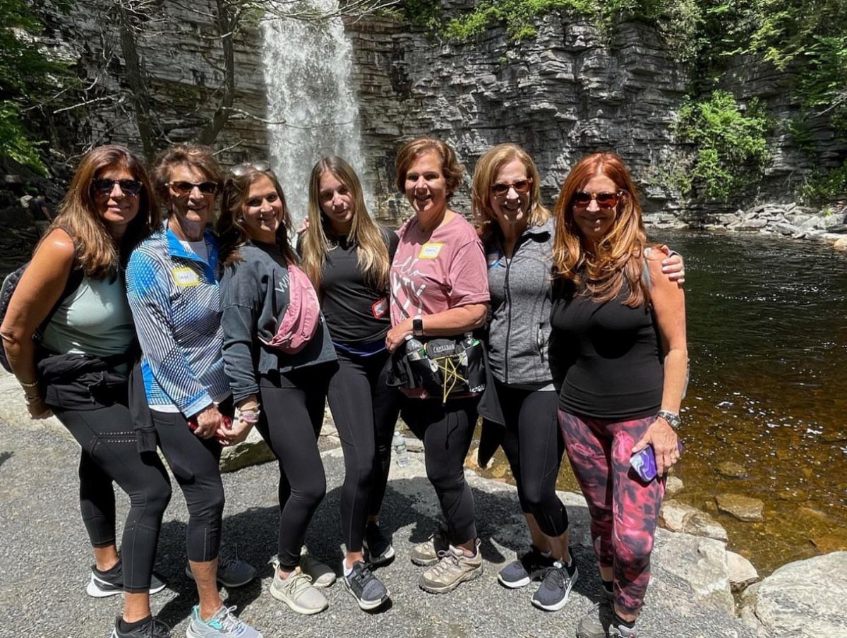 Seven women smiling together in front of a waterfall during an outdoor excursion.