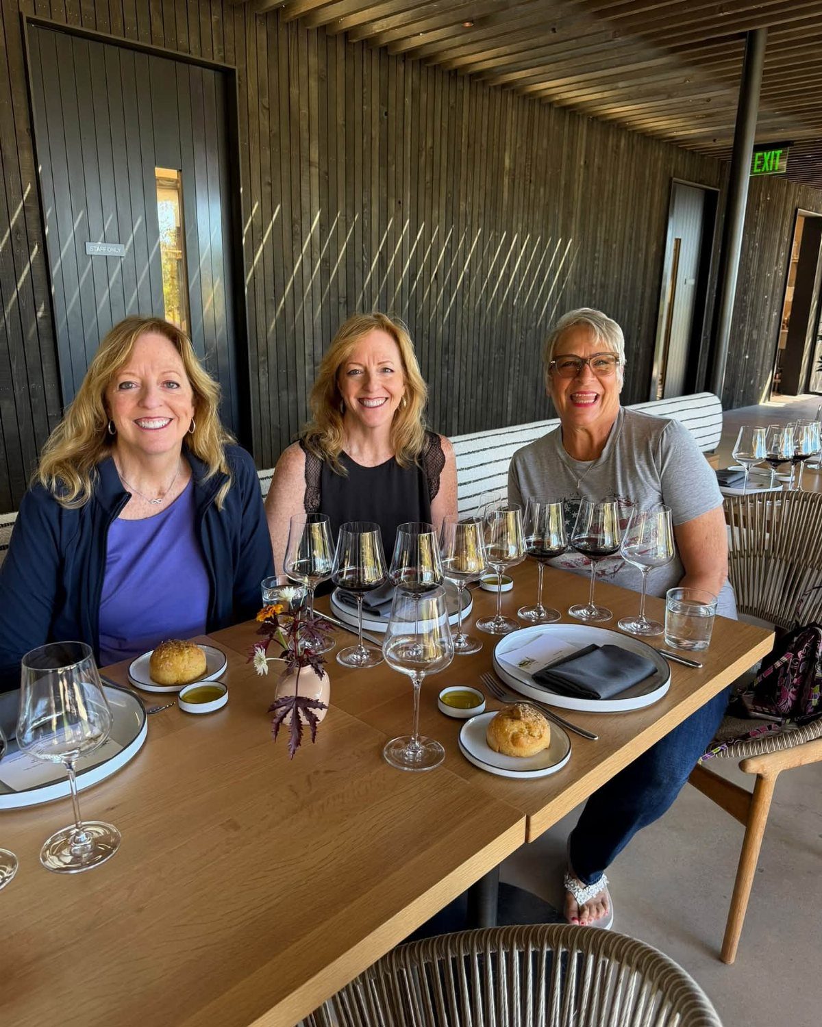 Three women smiling at a wooden table with wine glasses and pastries outdoors.