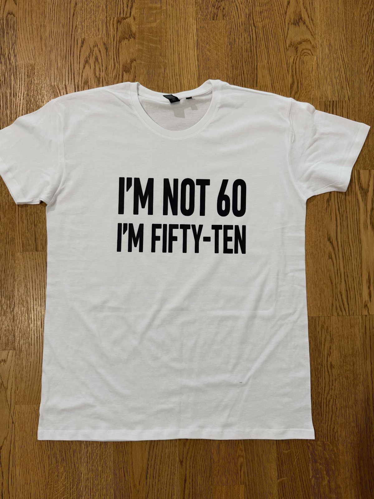 White t-shirt with black text saying I'm not 60 I'm fifty-ten on wood.