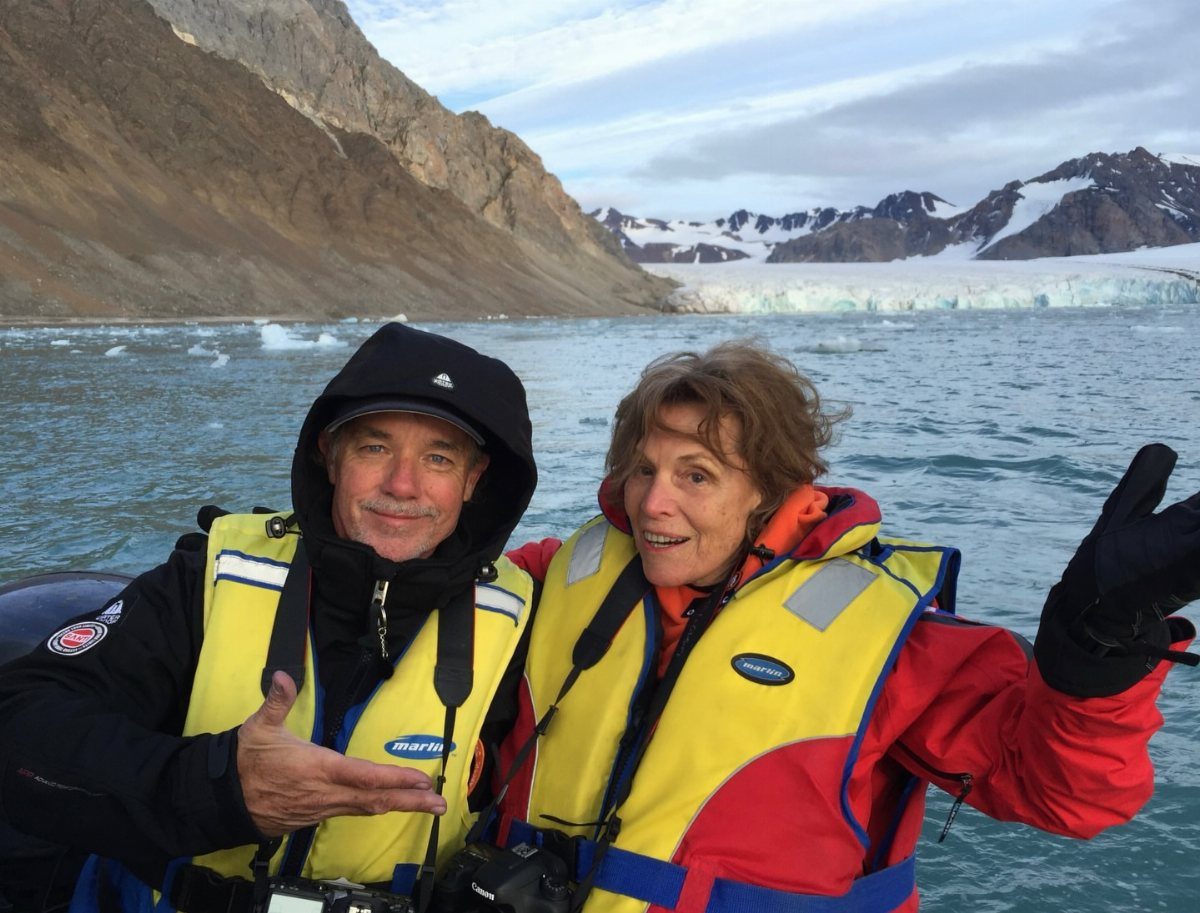 Older couple in life jackets posing on a boat before a glacier and mountains.