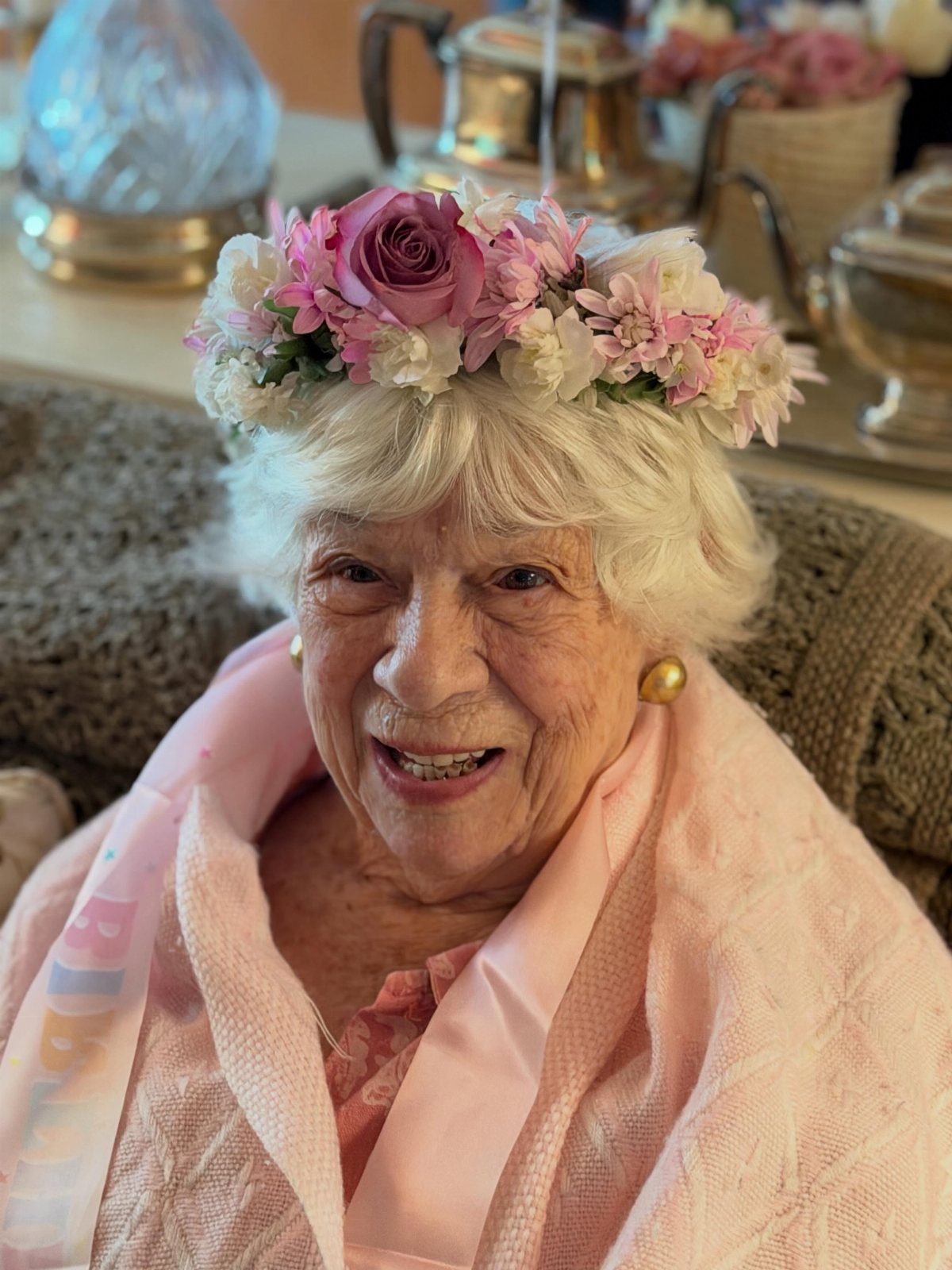 Elderly woman smiling while wearing a pink and white floral crown and shawl.