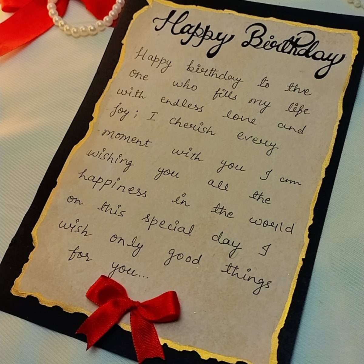Handwritten birthday card with burnt edges, a red bow, and a pearl necklace.