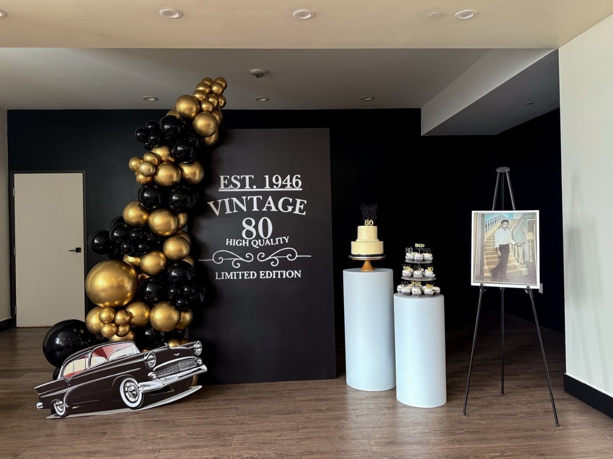 Birthday party setup with black and gold balloons, vintage car cutout, and 80th anniversary backdrop.