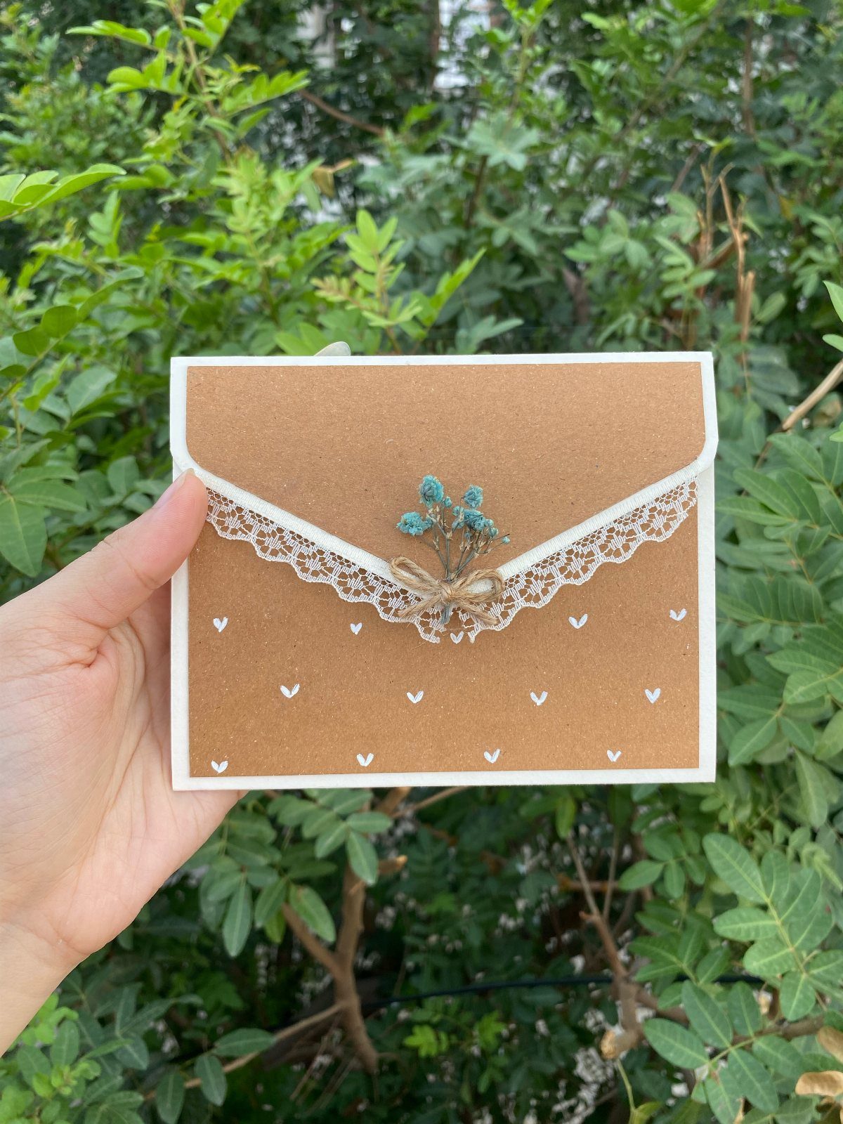 Brown kraft paper envelope with lace and dried flowers held against green leaves.