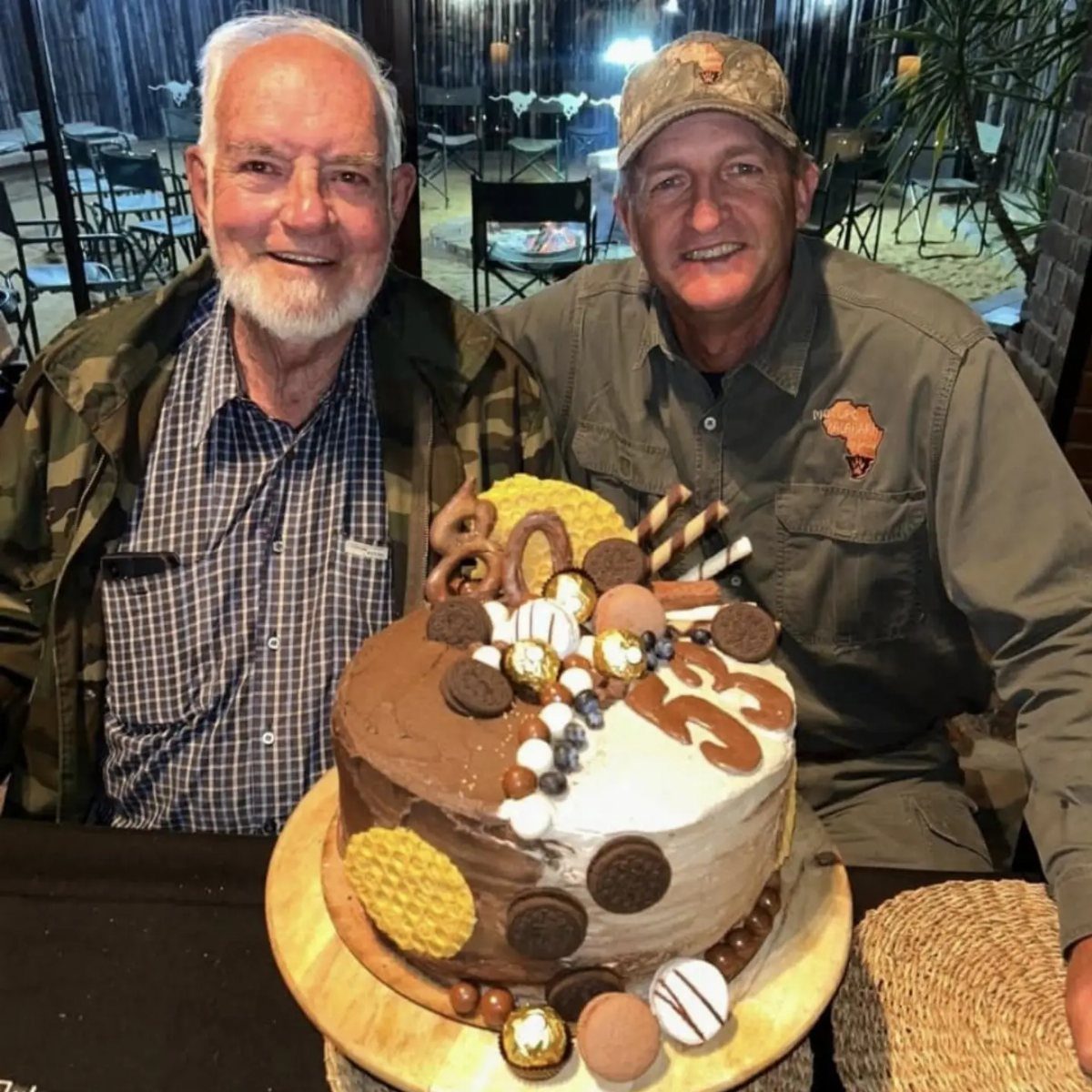 Two men smiling behind an 80th birthday cake in a safari setting.