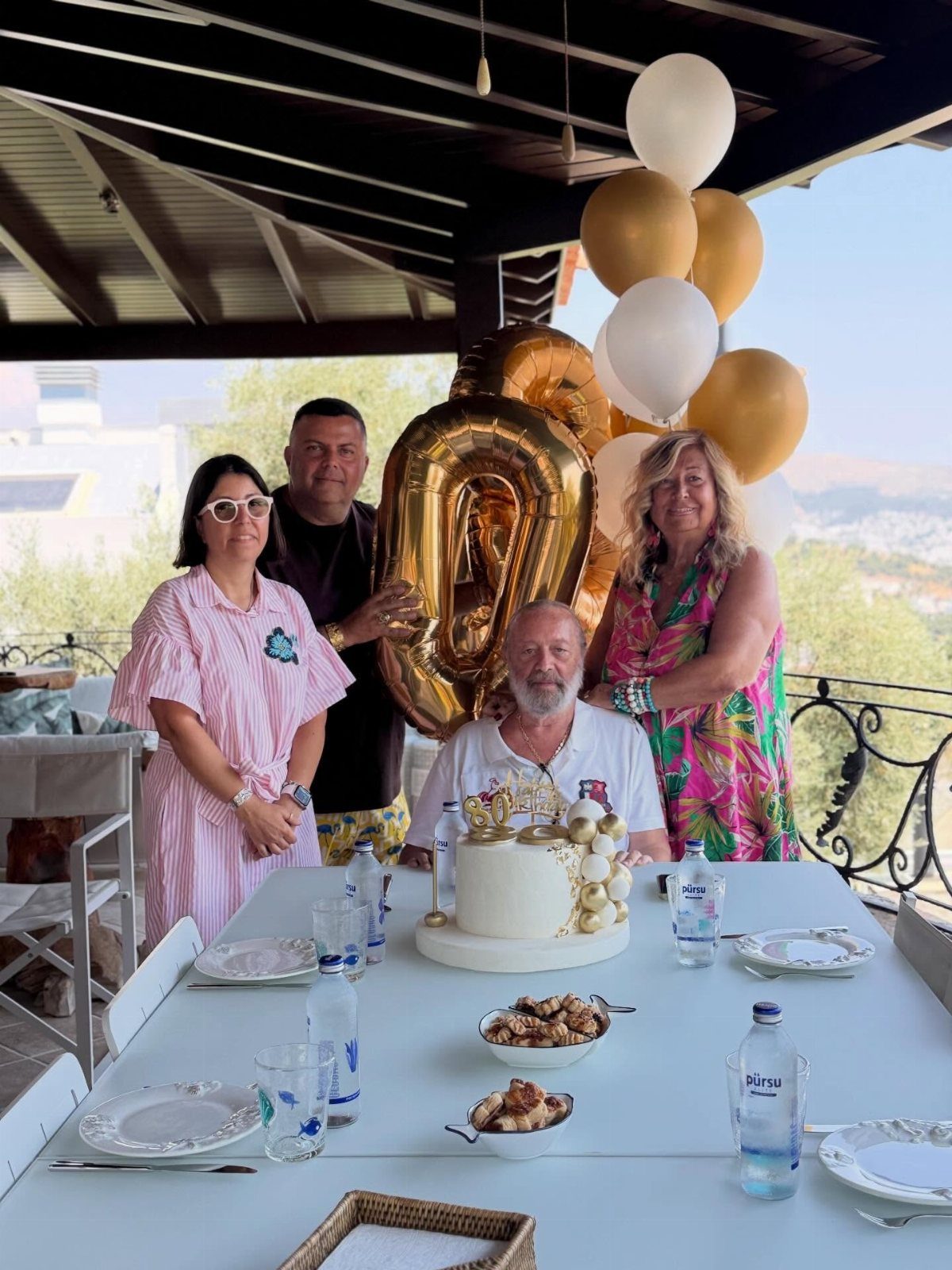 Family group posing around a birthday cake with gold balloons on a balcony.