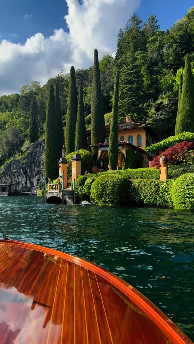 Wooden boat on blue water approaching a luxury villa with tall cypress trees.