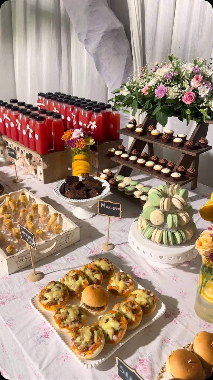 Buffet table with mini burgers, macarons, red drinks, and floral arrangements for tea.