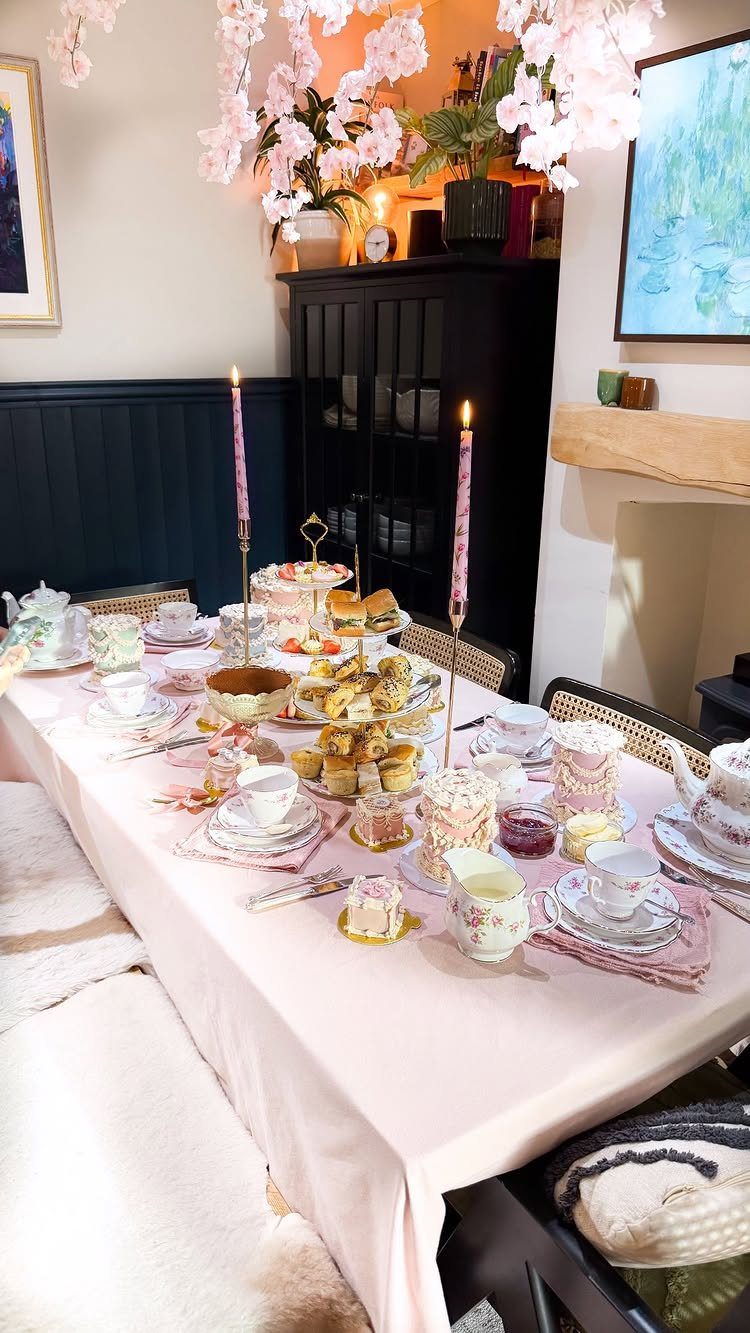 Afternoon tea table setting with pink tablecloth, tiered cake stands, and floral decorations.