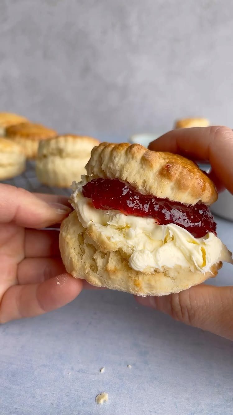 Scone filled with clotted cream and red jam held by two hands.