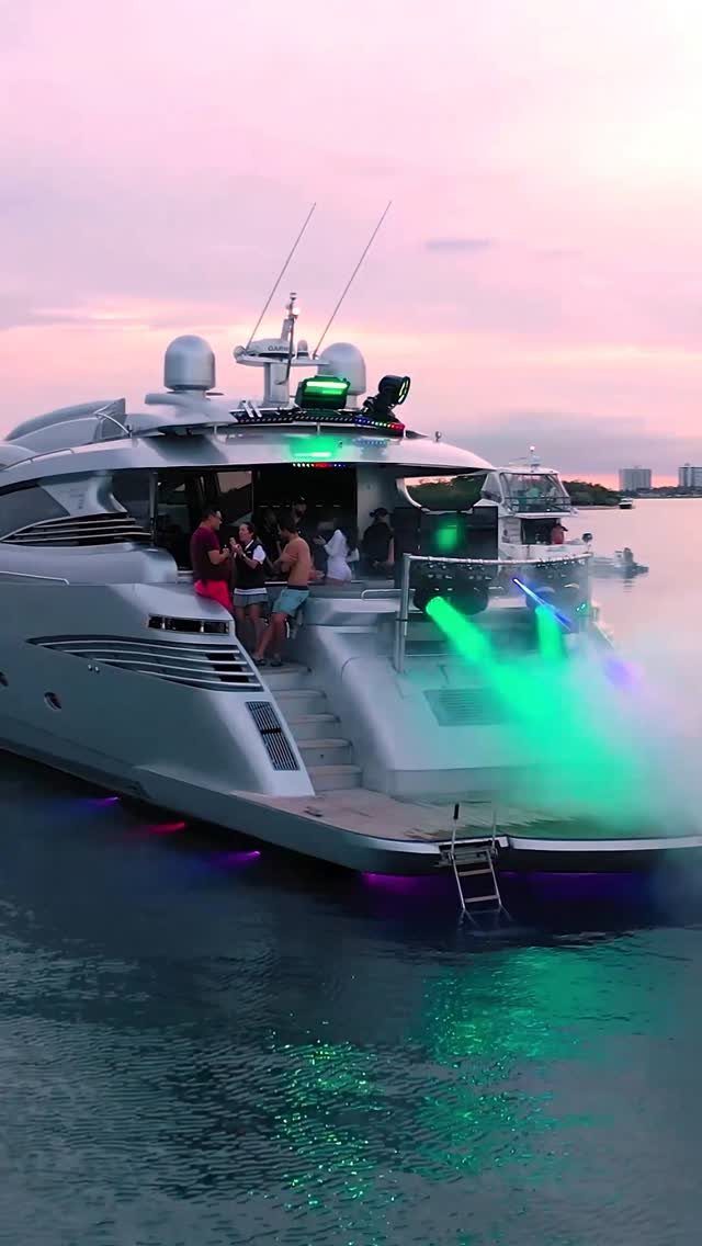 Modern silver yacht with party lights and people on deck at dusk.