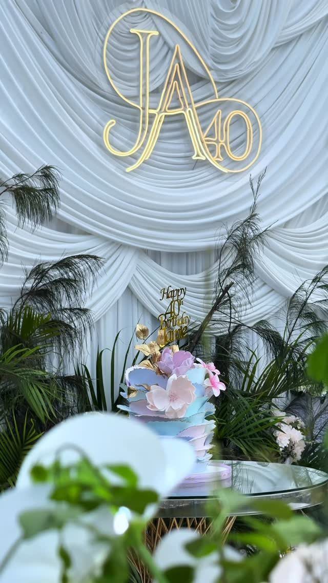 Tiered floral cake and gold 40th birthday neon sign against white draped fabric.