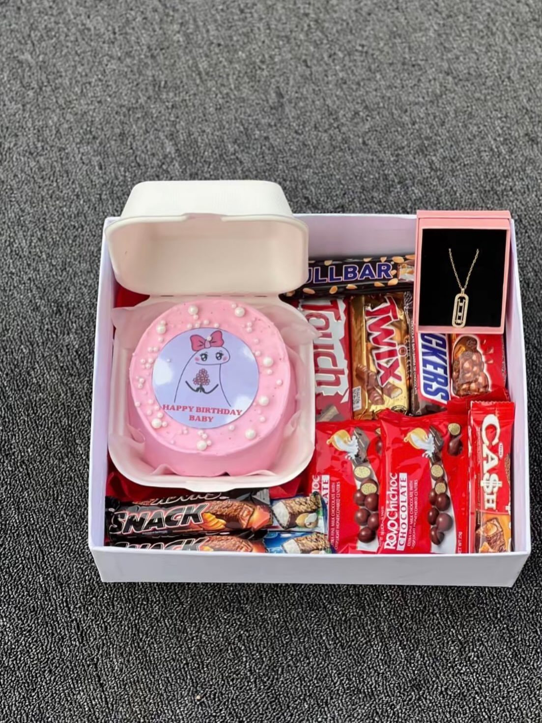 Pink birthday cake and assorted chocolates with a gold necklace in a gift box.