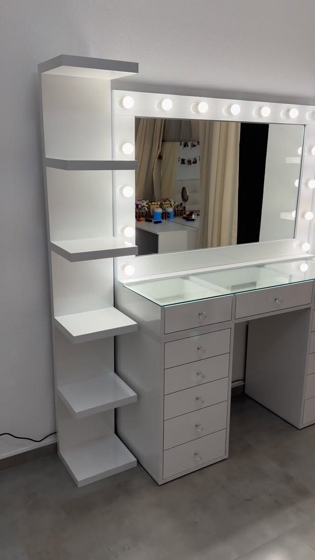 White vanity desk with a lighted mirror and side shelving unit against a wall.