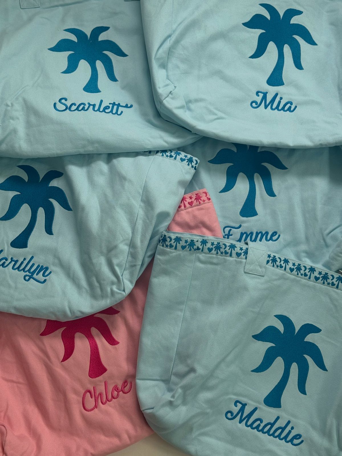 Personalized fabric tote bags with palm tree graphics and embroidered names in blue and pink.
