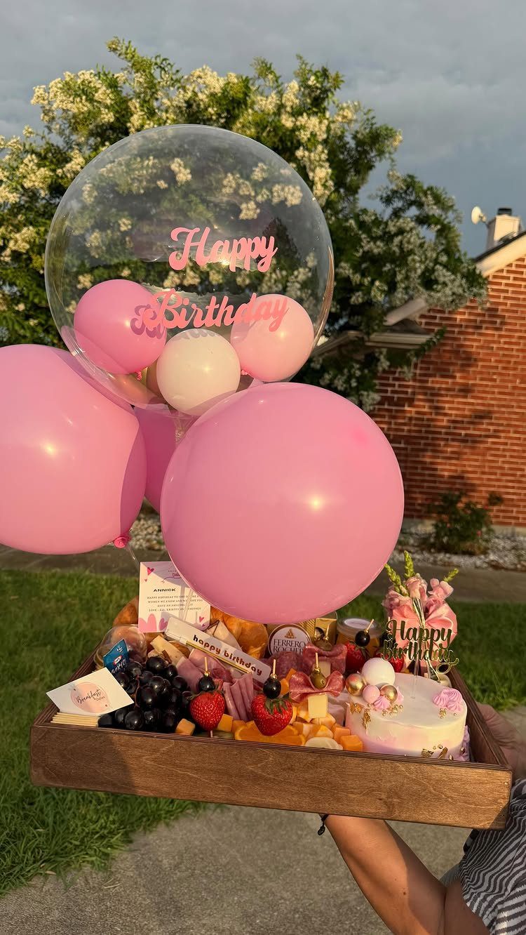 Birthday gift tray with charcuterie, a small cake, and pink balloons outdoors.