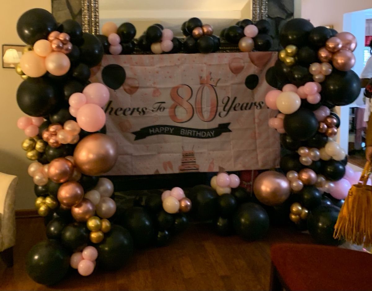 Rose gold and black balloon arch surrounding an 80th birthday banner indoors.