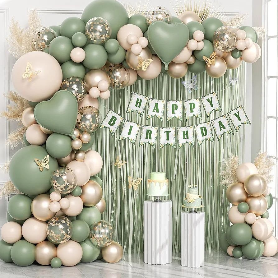 Sage green and gold balloon arch with birthday banner and cakes on pedestals.