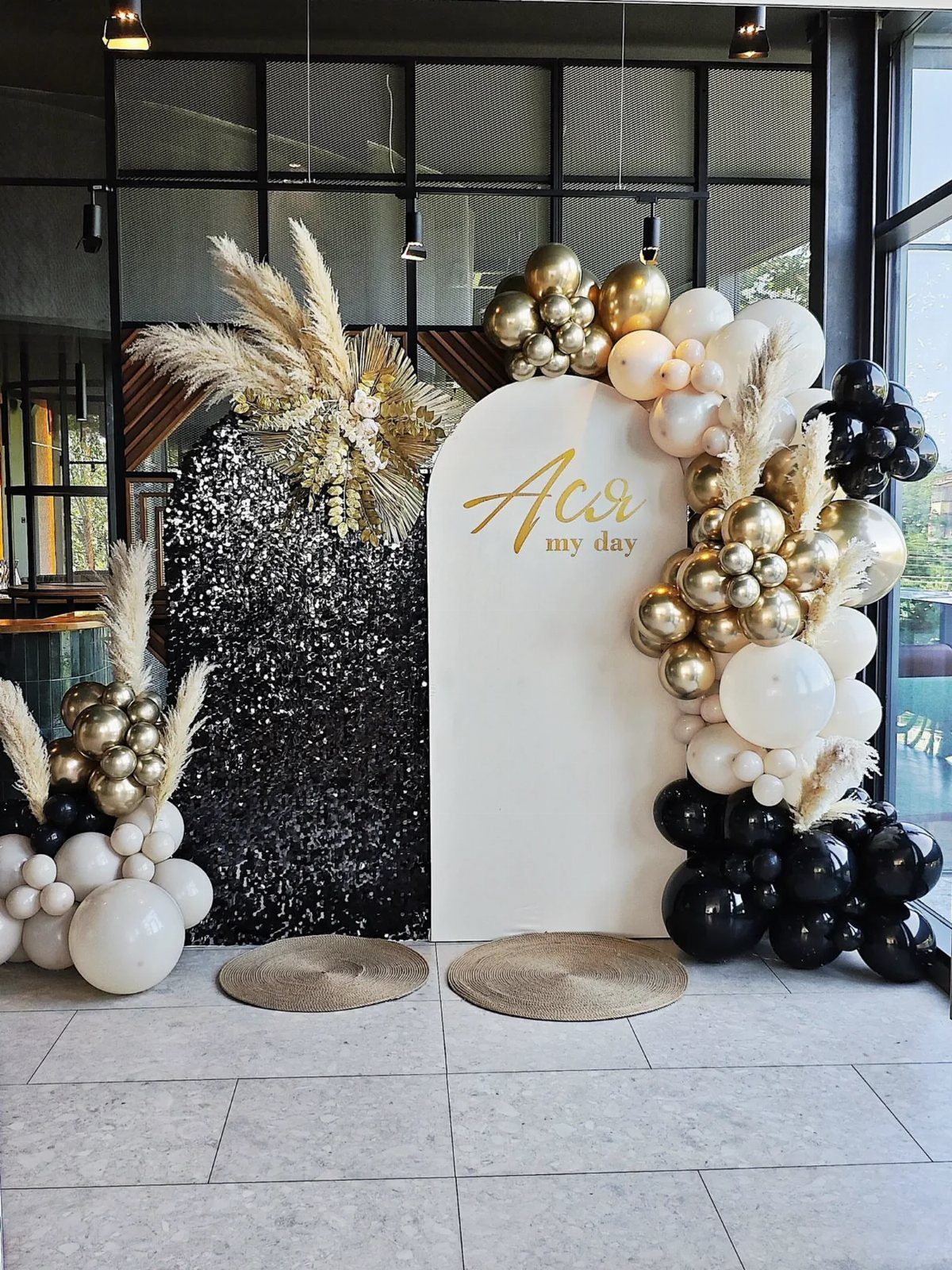 Party backdrop with black sequin wall, white arch, and gold, black, and white balloons.
