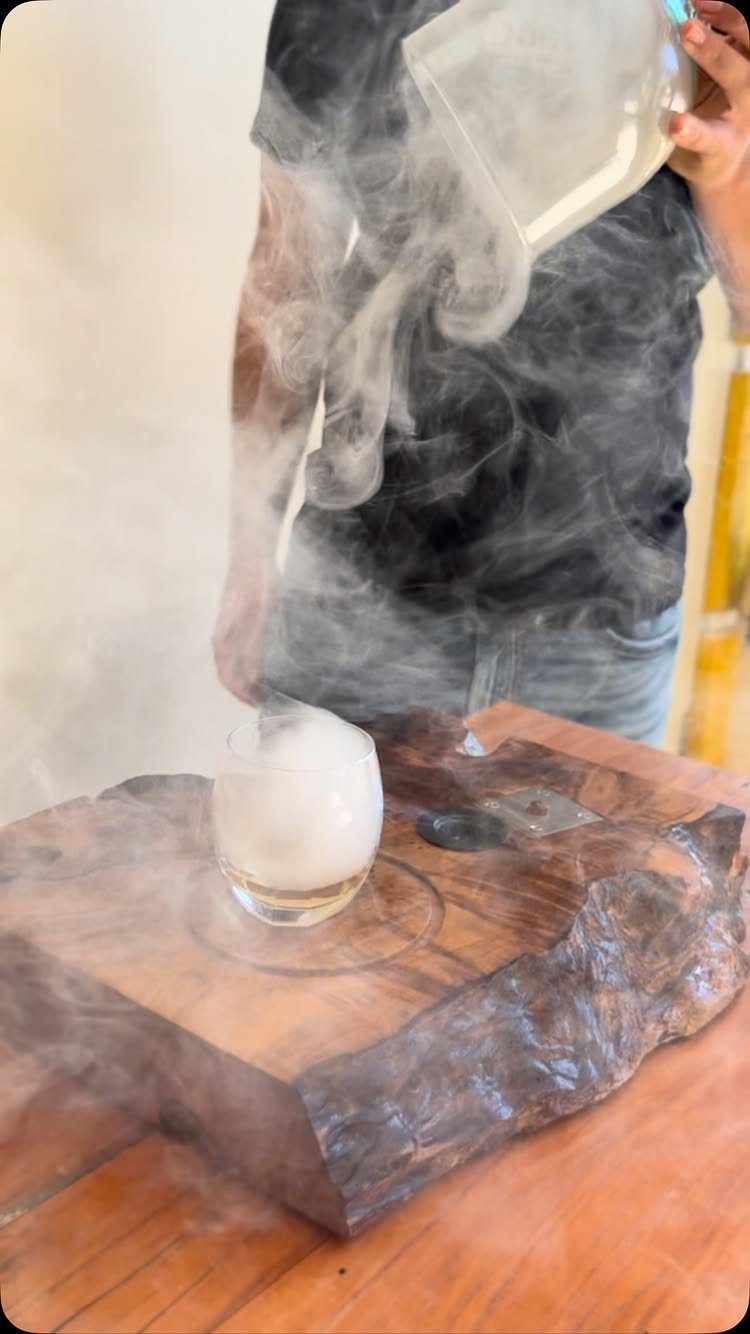 Smoky drink being poured into a glass on a rustic wooden slab