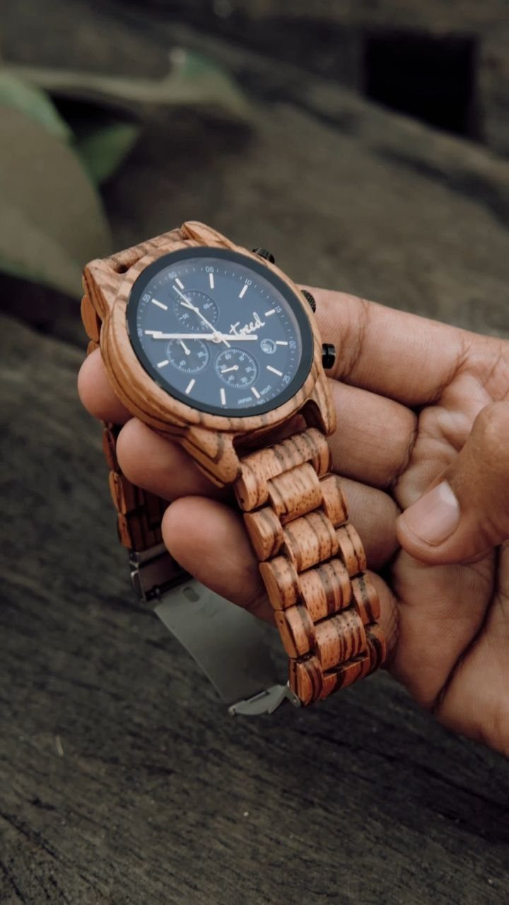 Wooden wristwatch with a dark face held in a person's hand over wood.