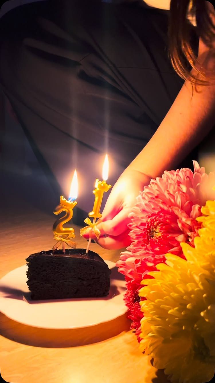 Chocolate cake slice with lit gold 21 birthday candles and colorful flowers.